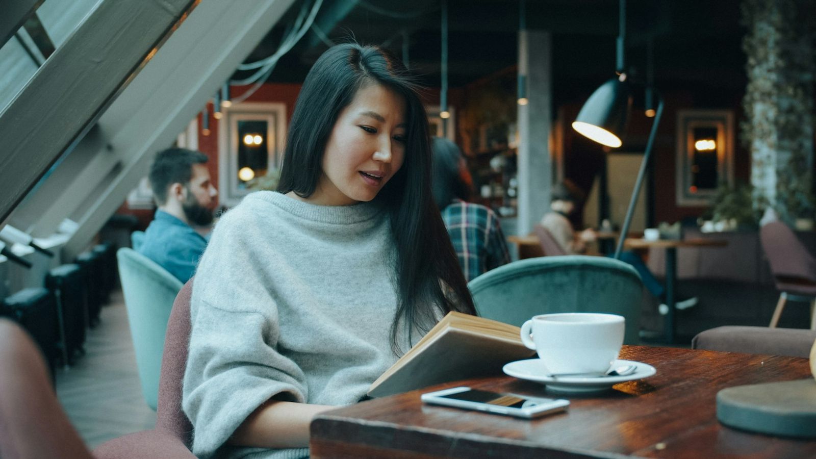 A woman reading a book at a cafe table, a cup of coffee and a phone beside her