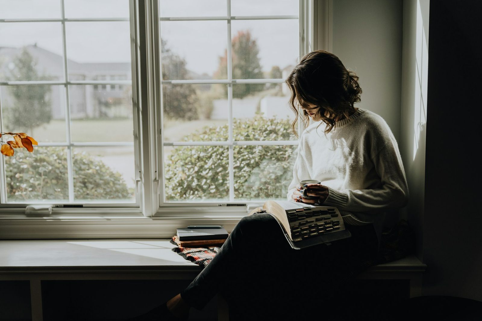 A woman reading a book by a window, holding a mug