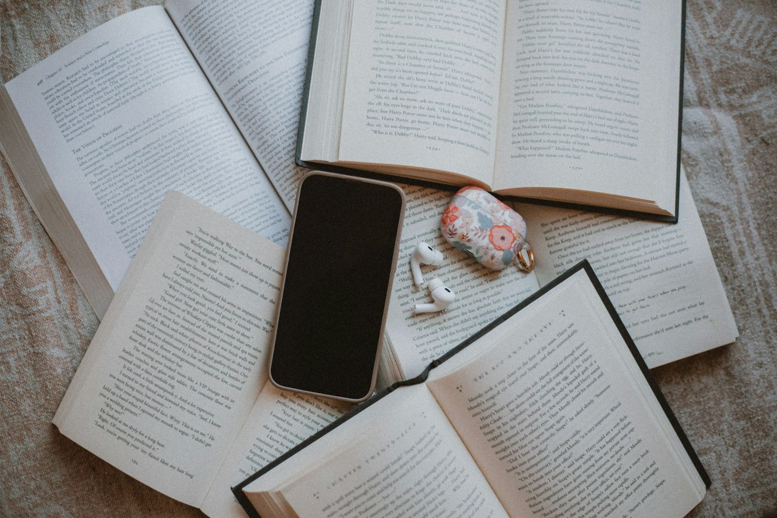 Open books on a bed with a phone and an earbuds case lying on the pages